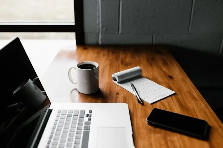 Laptop, coffee mug, notepad, pen, and smartphone on a wooden desk by a window, creating a cozy workspace.