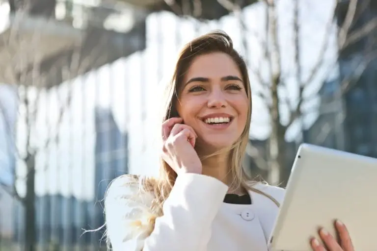 Smiling woman in a white coat holds a tablet and talks on the phone outdoors with modern buildings in the background.