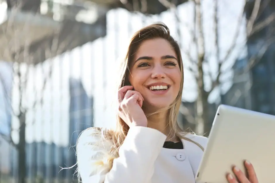 Smiling woman in a white coat holds a tablet and talks on the phone outdoors with modern buildings in the background.