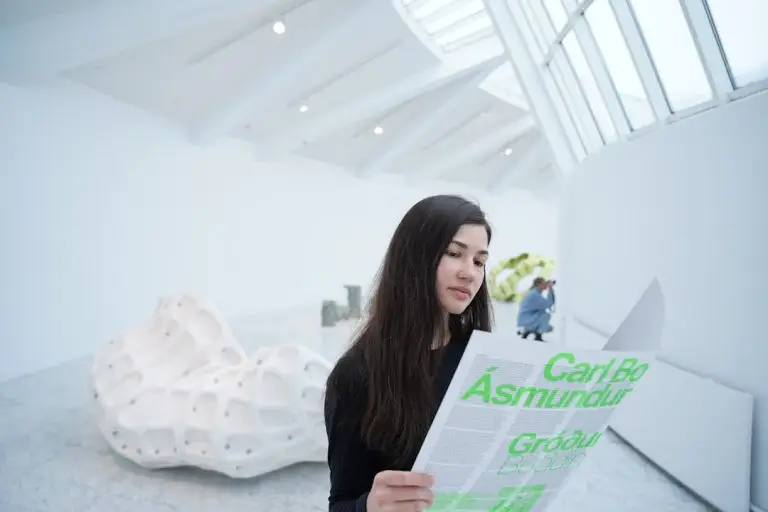 A woman in a modern art gallery reads a pamphlet, with abstract sculptures and a bright, skylit ceiling in the background.