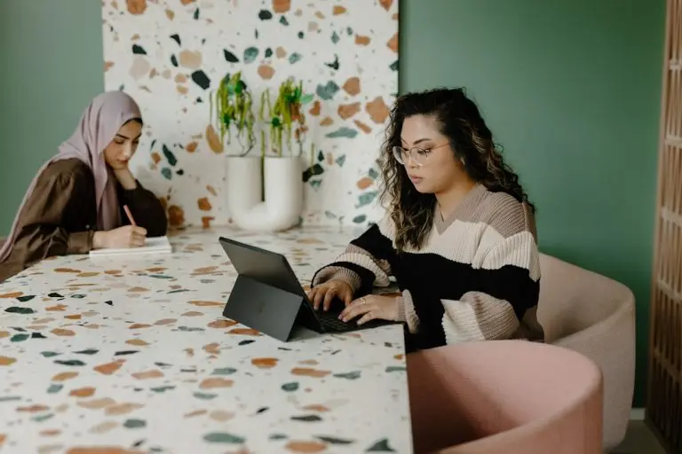 Two women working in a stylish office, one using a laptop and the other writing in a notebook at a terrazzo-patterned table.